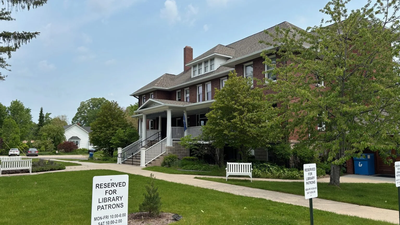 An old brick library with a wide white porch behind a lawn, trees and white benches, with 'Reserved for library patrons' parking signs in the foreground.