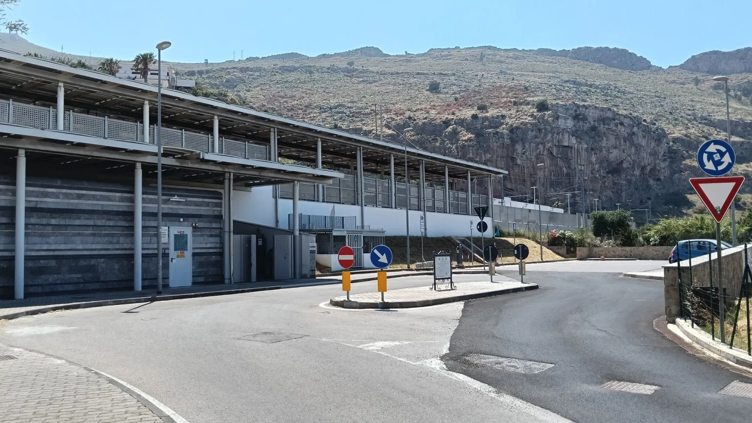 A small station with a long covered walkway beside a quiet road and roundabout signs, under a dry, rocky hillside.