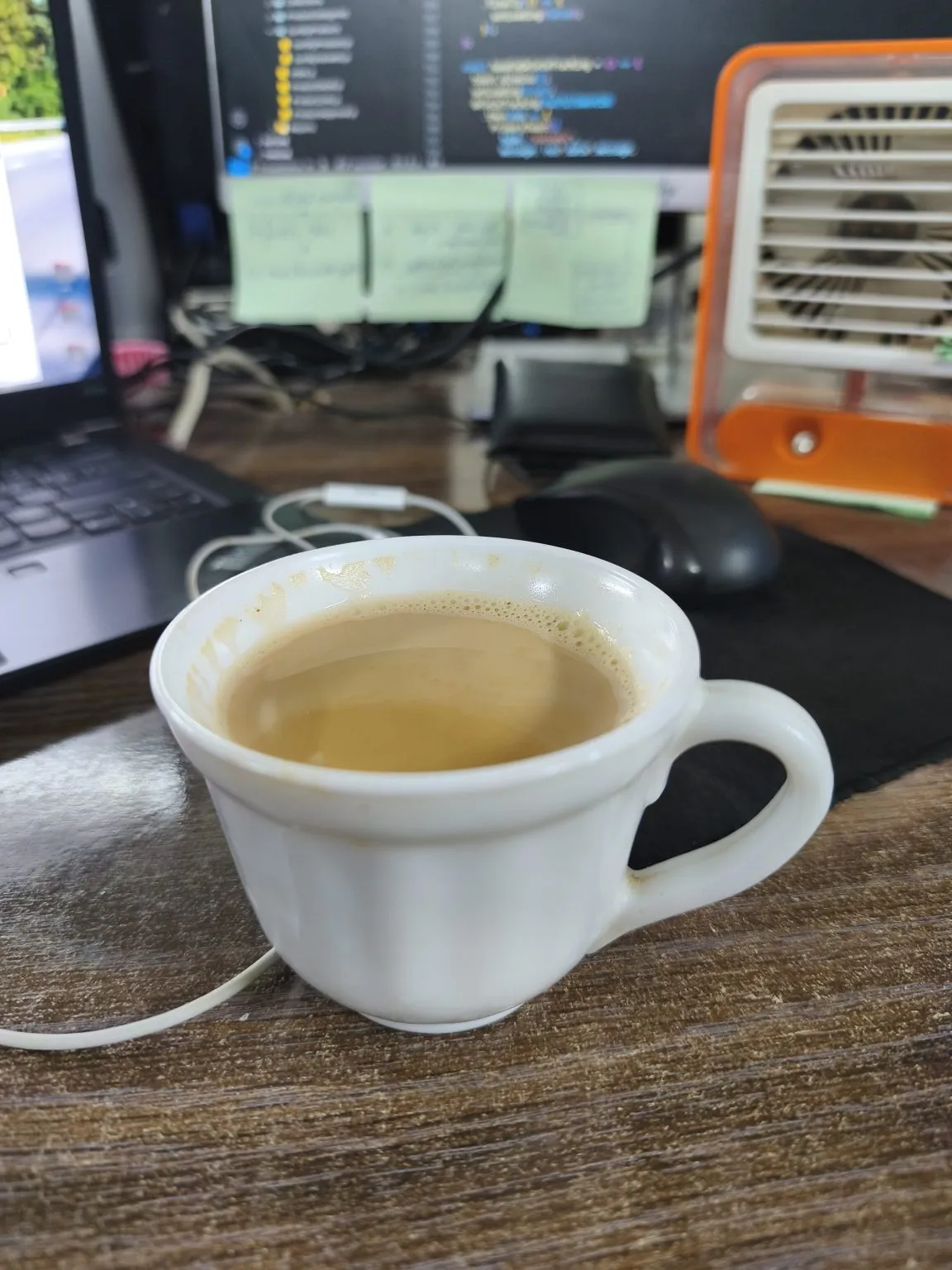 A white cup of milky tea on a wooden desk, with a laptop, three sticky notes on a monitor and a small orange fan behind.